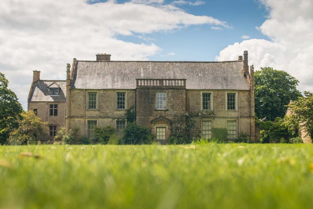 a large beige stone stately home with a grey tiled roof and tall windows in front of a grassy lawn