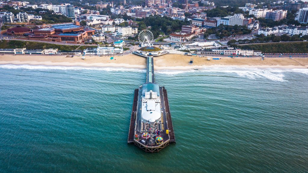 aerial shot of Bournemouth pier jutting out into the blue sea with the sandy beach on either side and part of the town behind. there is a large building on the pier and a white ferris wheel on the mainland just behind the beach. Things to do in Bournemouth Dorset. 