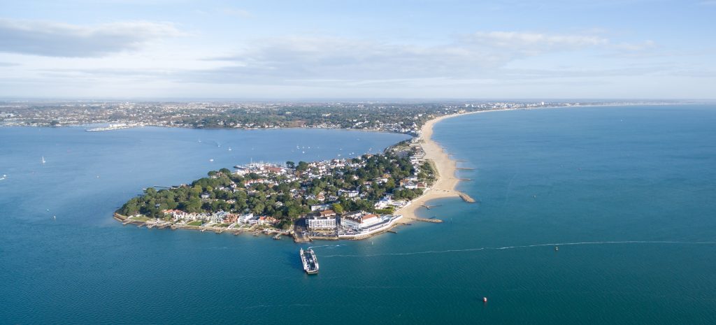 Aerial shot of Sandbanks peninsular between Poole and Bournemout, a small island with a leafy centre surrounded by a white sand beach, attached to the mainland by a thin strip of land with a long white sandy beach, all surrounded by bright blue sea