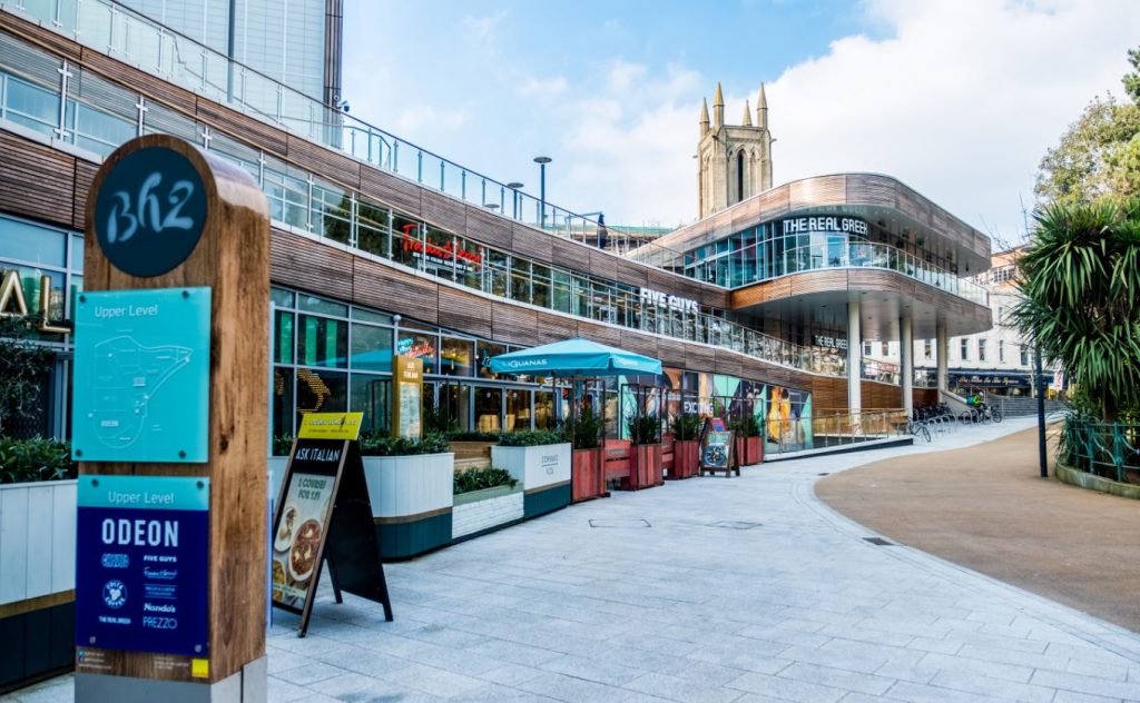 Exterior of the BH2 shopping centre in Bournemouth with a large pedestian paved walkway in front of several different restaurants on the ground floor level, the building has light wooden pannelling around the top of each floor and glass barriers around the upper floor walkways.