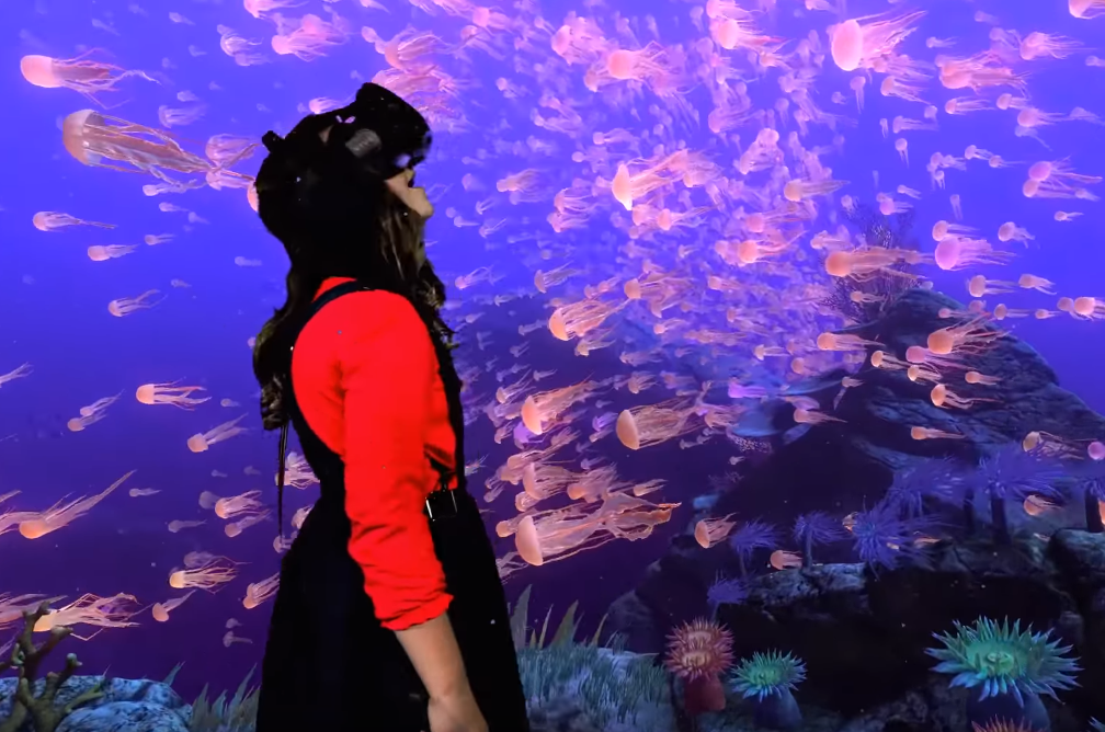 Woman with long brown hair wearing a red long sleeved shirt under a black dungaree dress and a virtual reality headset at Centre VR in Bournemouth, she is looking up with her mouth open as if in wonder, all around her is a projection of an underwater scene with a swarm of jellyfish swimming above a coral reef. 
