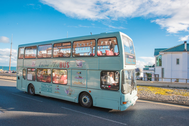 Doubledecker bus painted in pastel turquoise with the name Frieda's Tea Bus painted on the side, driving along on the seafront outside a white two storey building. Afternoon Tea Bus in Bournemouth