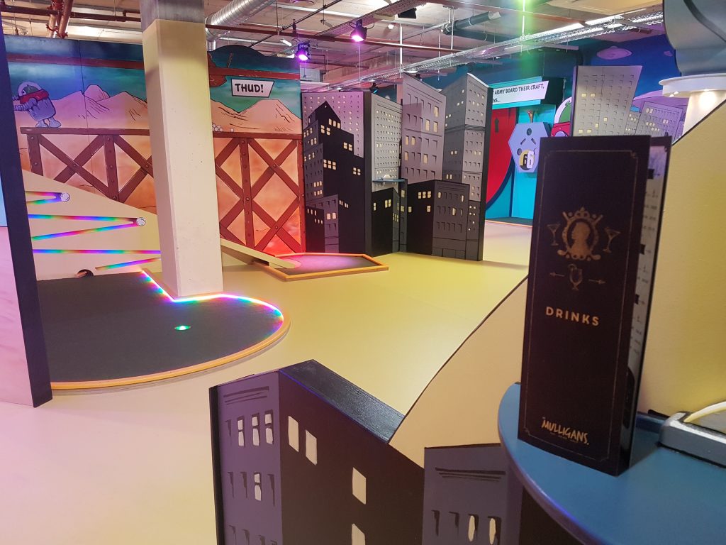 Interior of Mr Mulligans Indoor Adventure Golf in Bournemouth with a golf hole surrounde dby multicoloured neon lighting, all around it are large murals depicting a 2d cartoon city scape