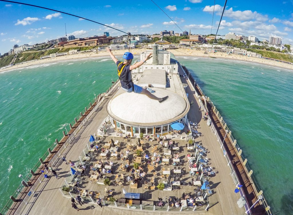 a man in shorts and a grey t shir with a blue helmet on is hanging from a zip wire above a pier with the sea on either side and bournemouth beach in the background. he has both his arms stretched out on either side. 