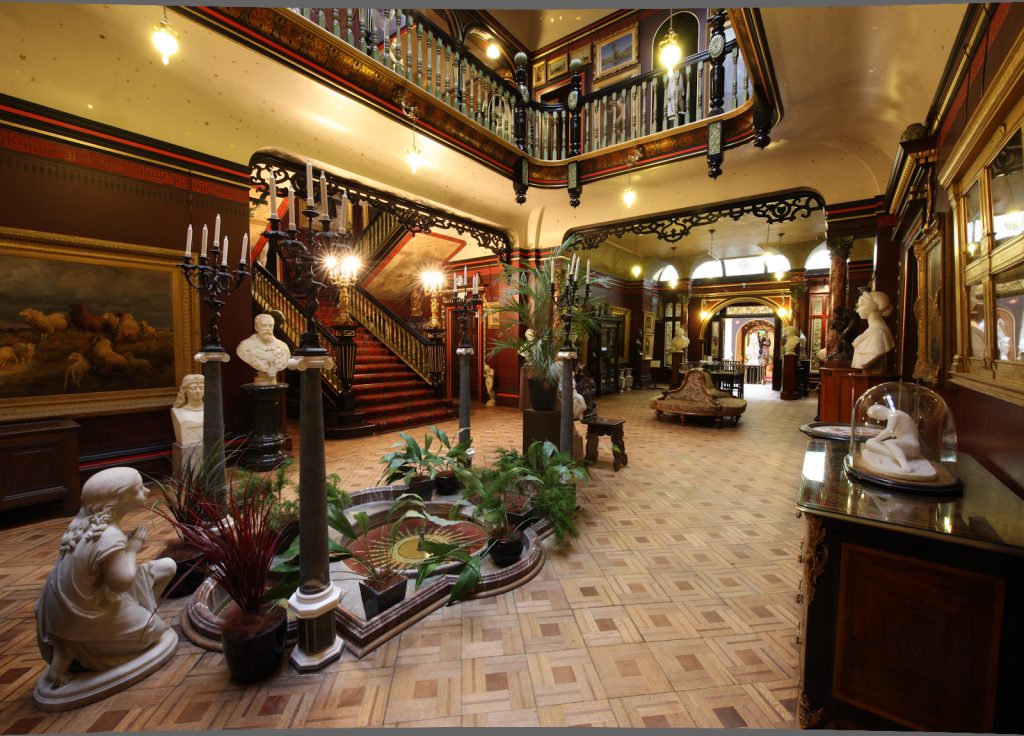 Interior of the Russell-Cotes Art Gallery and Museum with a large central hall with a floor made up of diamond shaped wooden tiles and cream walls with dark wooden trim and dark wooden railings around the uppper floor. there are many green houseplants and white marble statues around the hallway with antrique wooden furnishings around and several historic paintings on the wall. on the far side of the hall is a large double staircase with dark wood and a dark red carpet. 