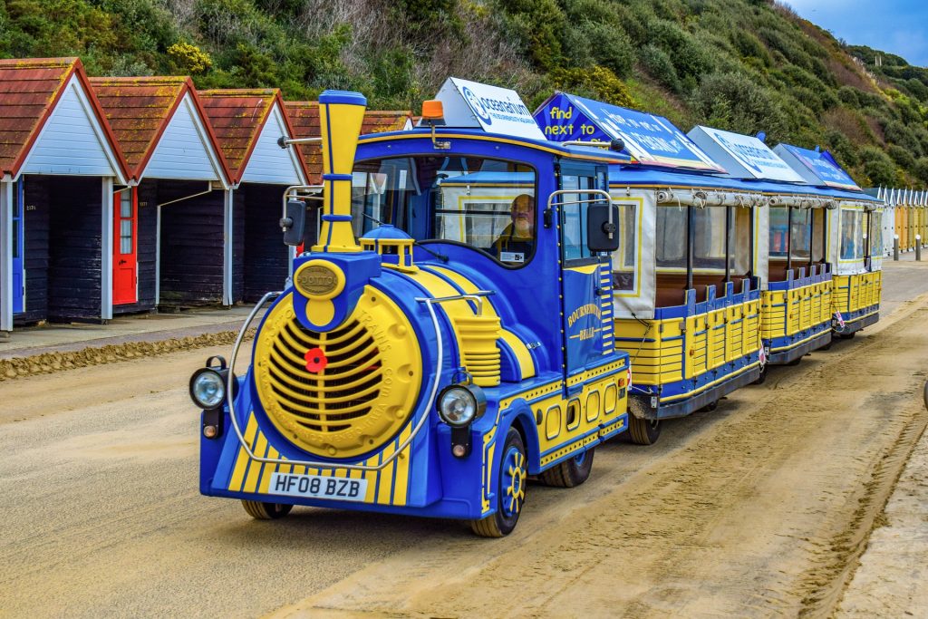 Blue and yellow land train running along a paved esplanade covered in a lot of yellow sand with a line of white beach huts behind with red tiled roofs and a sloping cliff edge covered in green shrubbery and long grass behind those. 