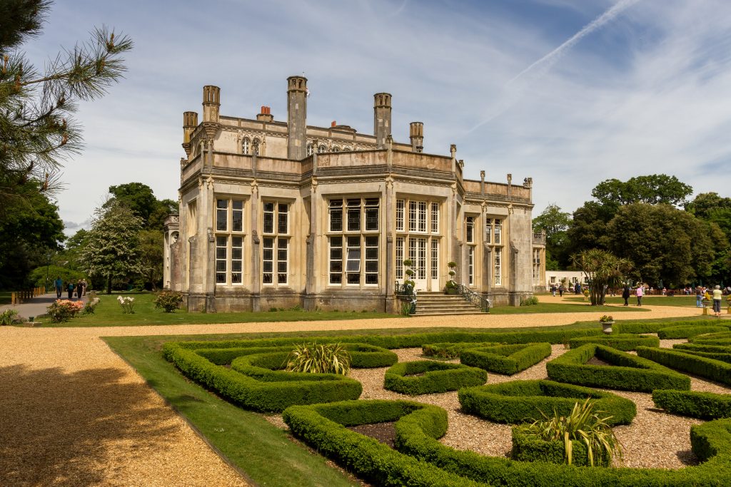 exterior of a small castle built from beige stone with a miniature hedge maze in front and a gravel path around it on a sunny day with blue sky behind. highcliffe Castle in christchurch near bournemouth. 