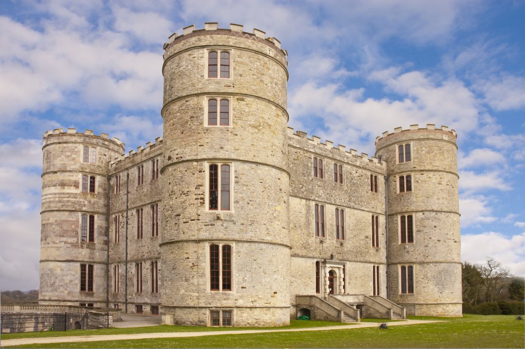 Exterior of Lulworth Castle in Dorset, a grey stone castle in a square shape with a round turret on all four corners, surrounded by a grassy lawn on a sunny day with blue sky above