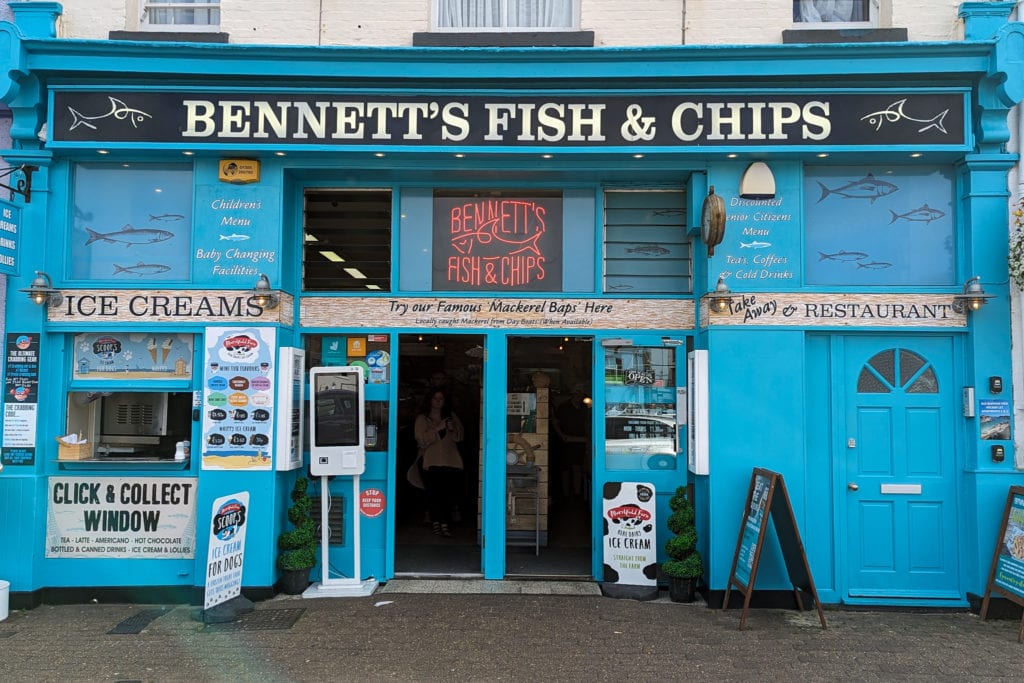 Exterior of Bennets Fish and Chips Weymouth, the building is painted bright cyan with a black and white sign displaying the shop name along the top and a small ordering hatch to the left of the entrance door