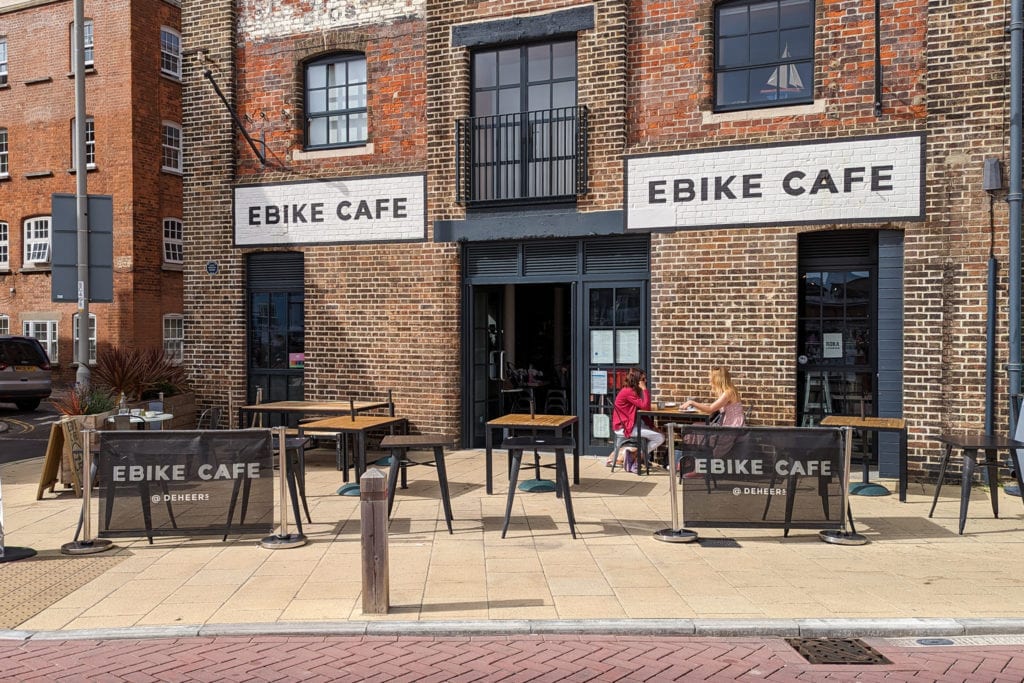 exterior of a red brick building with tables on the yello wpaved terrace outside and two white stone signs on the building reading "ebike cafe"