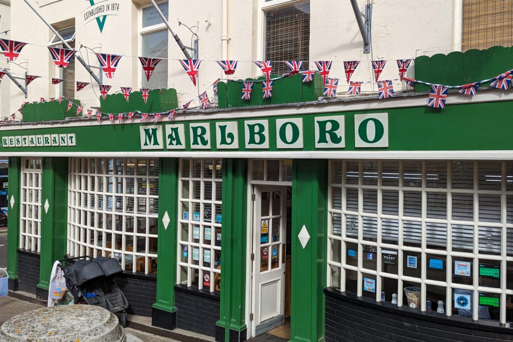 exterior of the Marlboro Fish and Chips shop in Weymouth with a green painted facade and long pannelled windows and the shop name printed in green letters on cream squares along the top beneath some union jack flag bunting