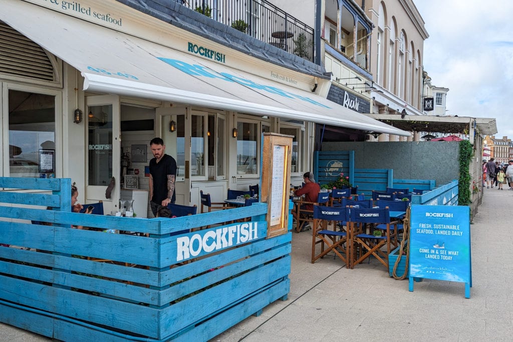 Exterior of the Rockfish restaurant on Weymouth esplanade with a bright blue fence around the outside seating area and a large cream awning above with the logo printed in cyan