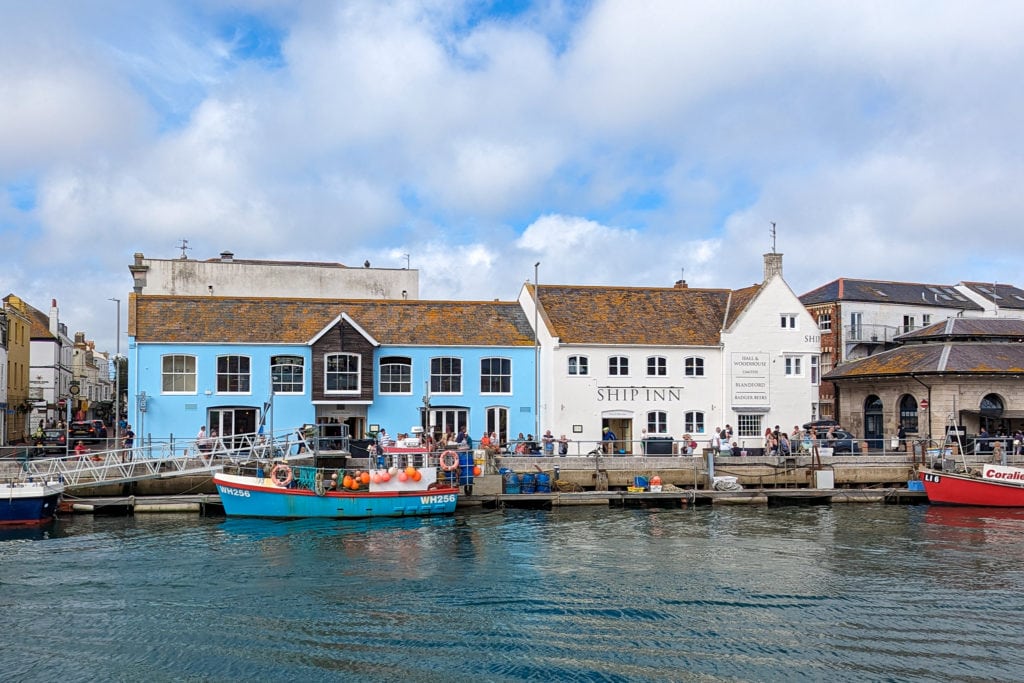 large pub with one cyan building and one white on the harbourside in weymouth viewed from across the water with two wooden fishing boats, one blue and one red, moored in front and a cloudy blue sky overhead on a sunny day. Best places to eat in Weymouth. 