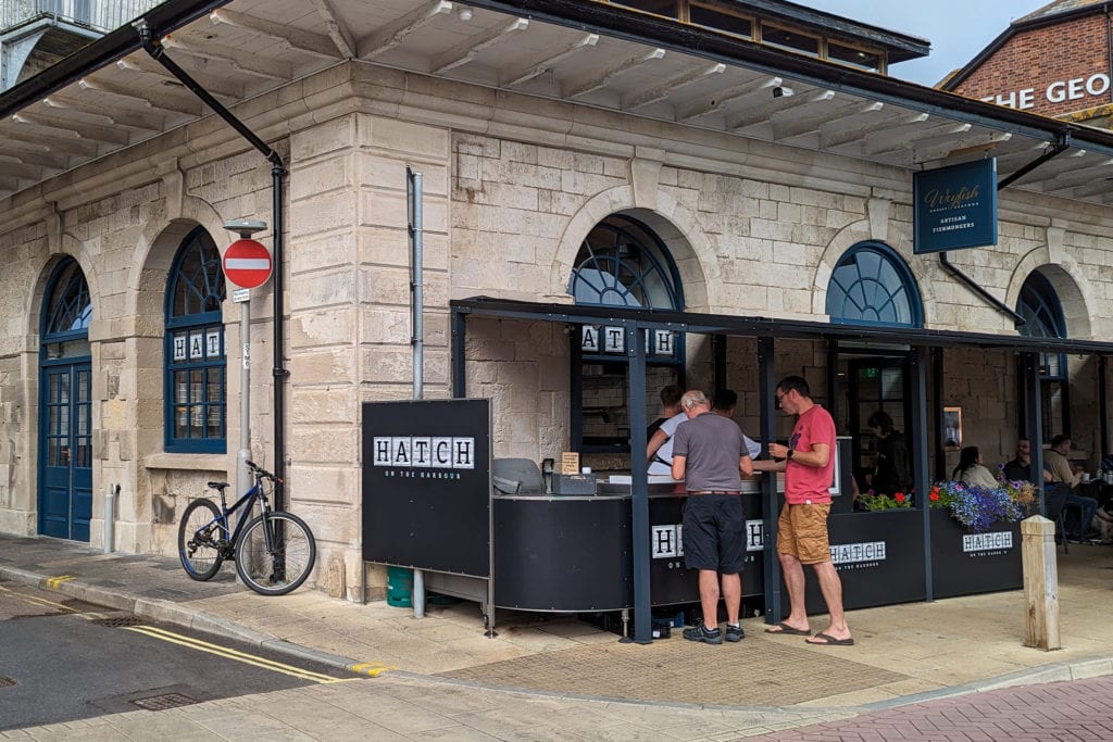 two men ordering from a black painted street food stall on the corner of a one storey stone building 