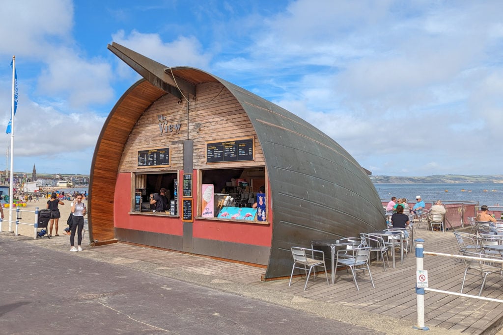 semi circular domed food hut with a wood panneled front with two hatches for ordering and a grey wood pannelled domed roof, on the side of the esplanade next to weymouth beach with an outdoor dining area to the right of the hut
