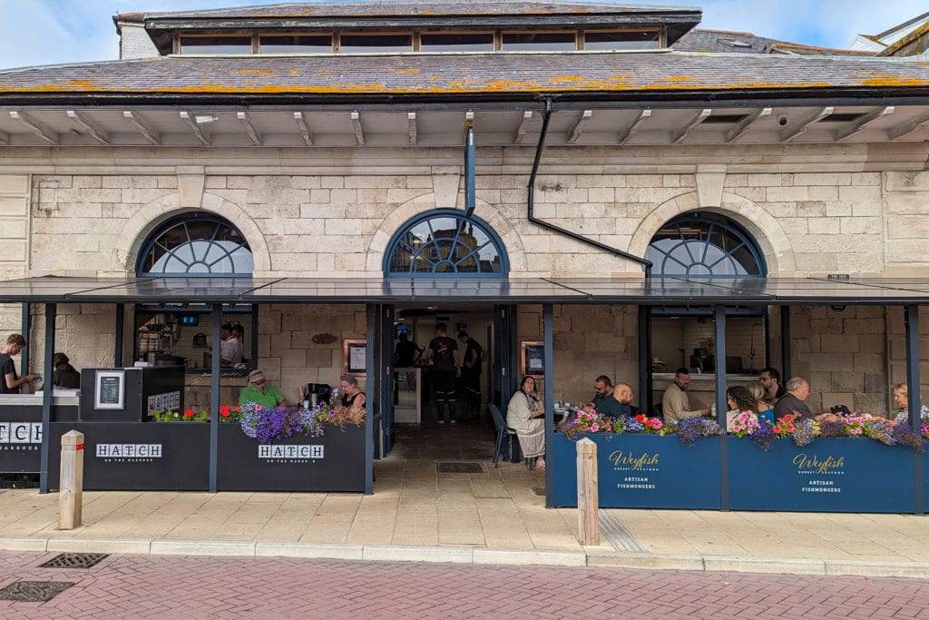 one storey stone building with arched windows with restaurant tables outside behind a blue banner with the Weyfish fishmongers logo on it, on the left of the building is a food stall with black banners and the name Hatch printed on it in white