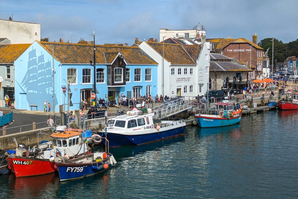 Blue and white buildings along the edge of Weymouth Harbour with a line of fishing boats moored along the harbour wall on a sunny day with cloudy blue sky above. Best Places to Eat in Weymouth. 