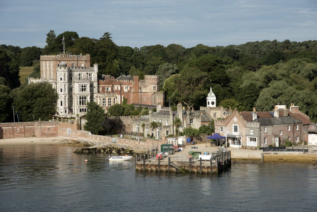 a castle built in a mix of grey stone and red brick with a flag on top. there is a forest behind and the sea in front with a small dock and a little white boat next to it. there is a tiny village of stone buildings behind the port with a white clocktower rising above it. 