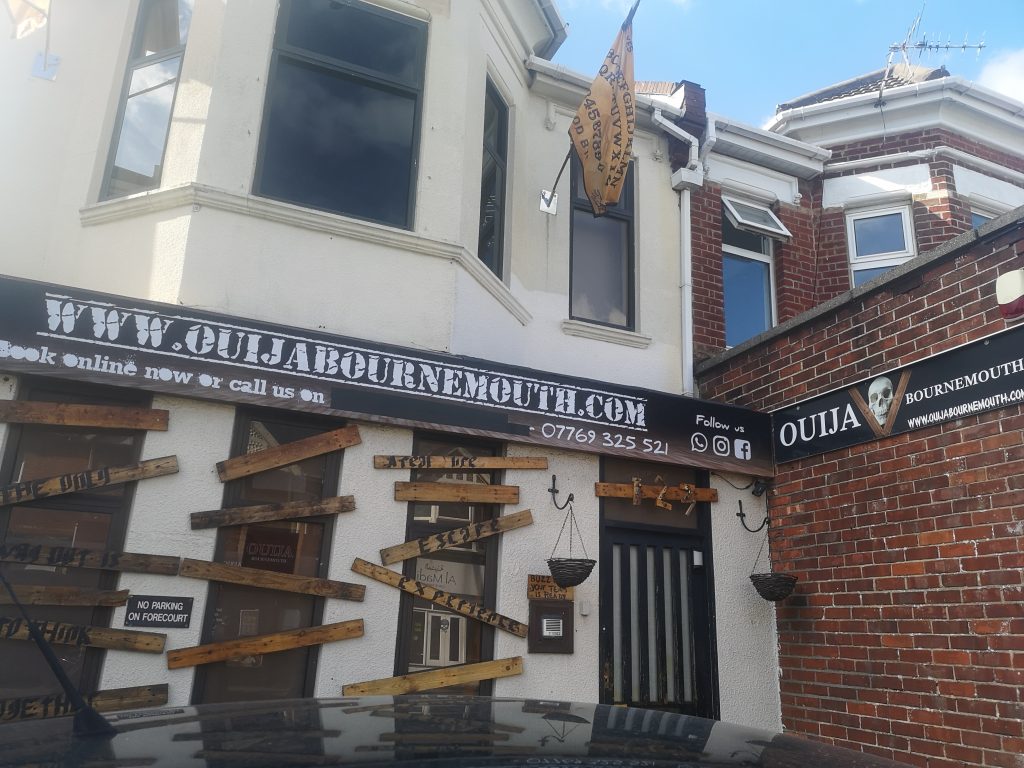 Exterior of the Ouija Escape Room in Bournemouth, a shop front with boarded up doors and windows on the lower half of a white building with a yellow flag between the second floor windows