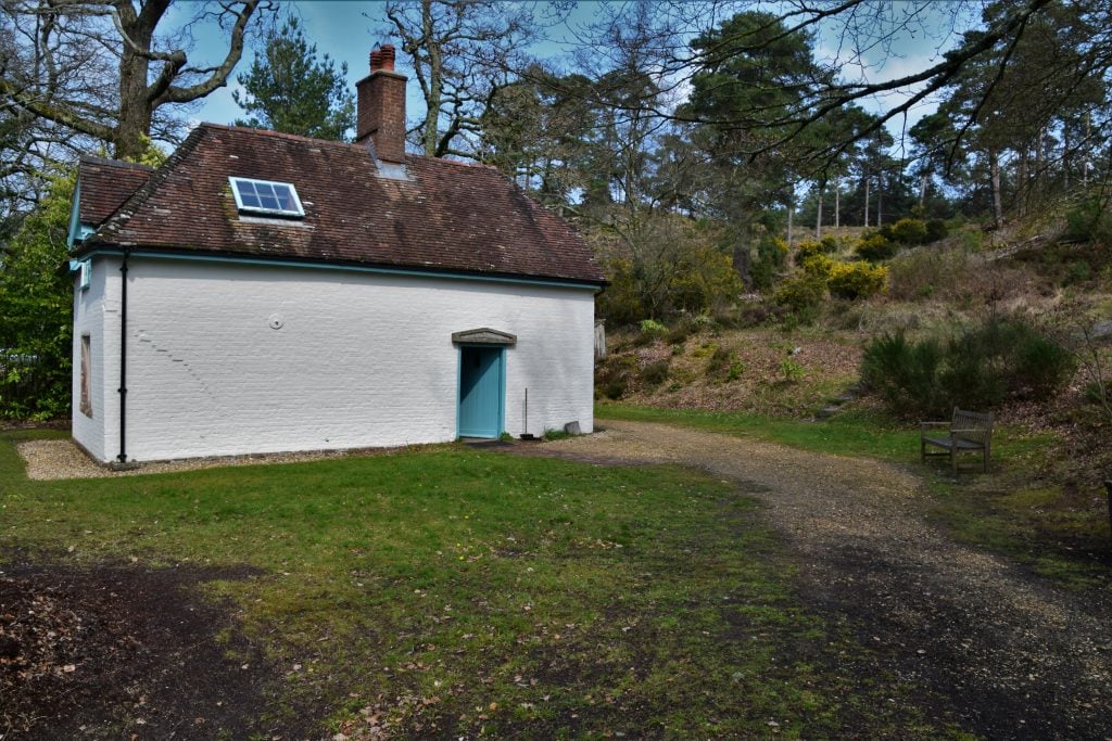 tiny white cottage with a blue door and a single window in the grey slate roof surroudned by trees with a lawn in front