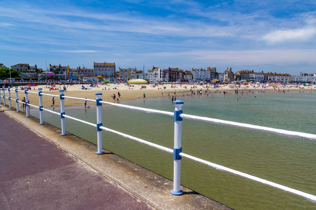 Looking through a painted white fence towards the sea with a sandy beach in the background and a row of houses and hotels on Weymouth seafront