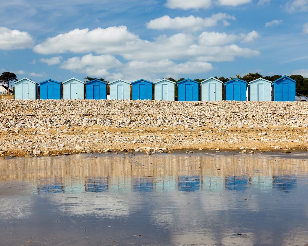 a row of beach huts painted in alternating shades of pastel and bright blue with a shingle beach in front all reflected on the calm sea on a sunny day - best things to do in charmouth