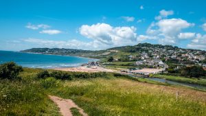 Panoramic view of Charmouth, a small seaside town sitting in a bay with bright blue sea, viewed from the top of a hill with a patch of grass in the foreground