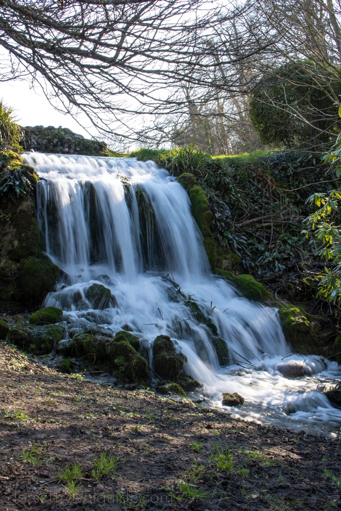 long exposure of a wide waterfall flowing quickly in two layers down a rock face with some bare winter tree branches overhead