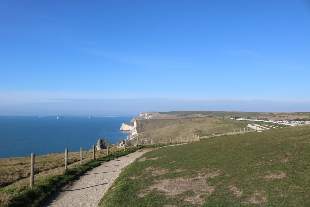 white gravel path at the edge of a grassy cliff with the sea below and white cliffs along the coast in front, taken on a sunny day with clear blue sky above