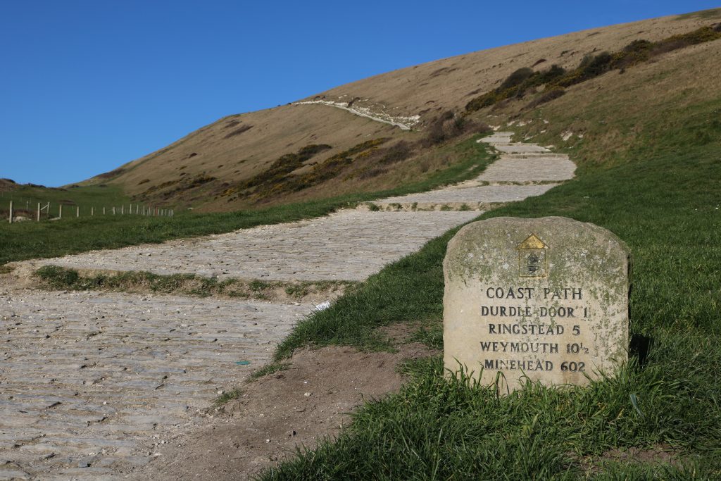 small stone signpost for the SW Coast Path next to a white gravel path leading uphill along a grassy slope with blue sky above