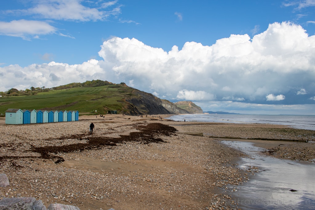 Shingle beach in Charmouth dorset with green cliffs behind and a row of blue beach huts to the left
