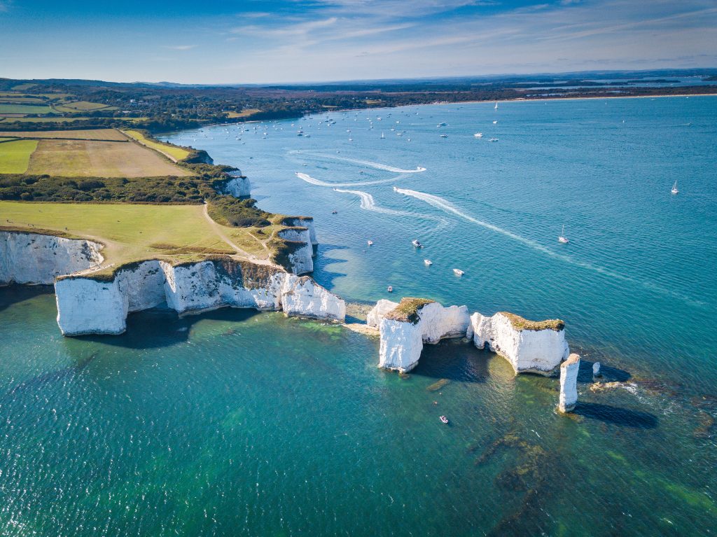 drone shot of a headland topped with grass with white cliffs and a thin white line of chalk rock and rock stacks stretching out into the bright blue sea on the isle of purbeck in dorset with the coastline in the background