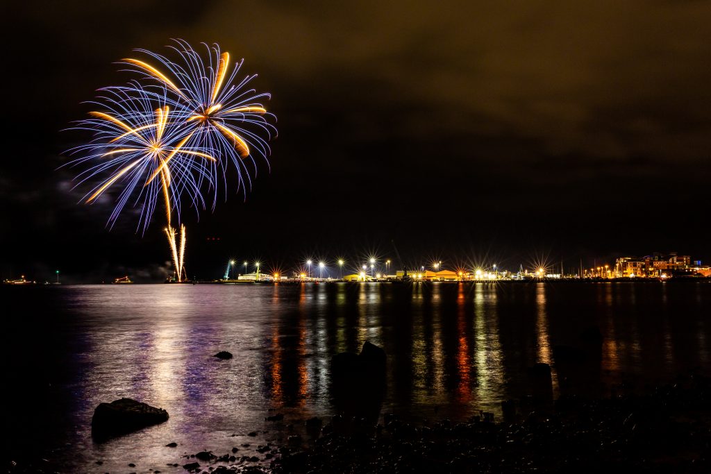 Poole harbour at night with fireworks exploring in the sky above the sea at the end of the quay, which is lined with buildings and lights. 