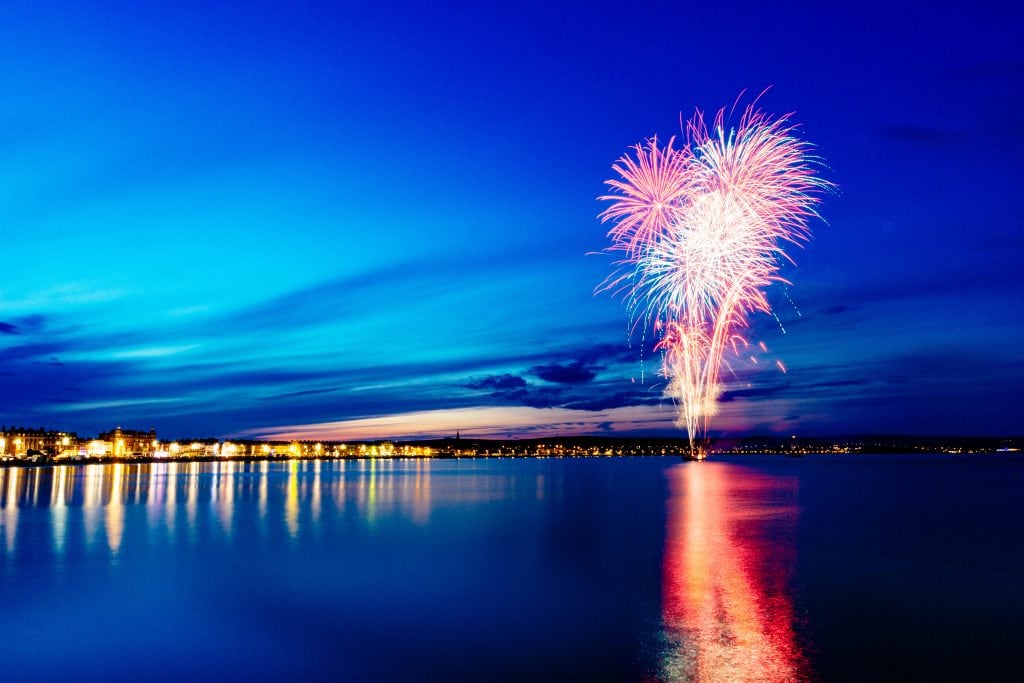 Fireworks exploding in the night sky above Weymouth Bay in Dorset, the explosions are red and yellow and reflecting in the blue sea, with the lights of the town lining the shore. 