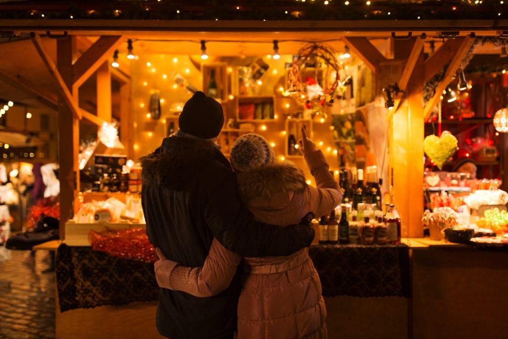 Couple in thick winter coats and wooly hats standing with their arms round each other in front of a market stall covered in fairy lights at night. Dorset Christmas Markets