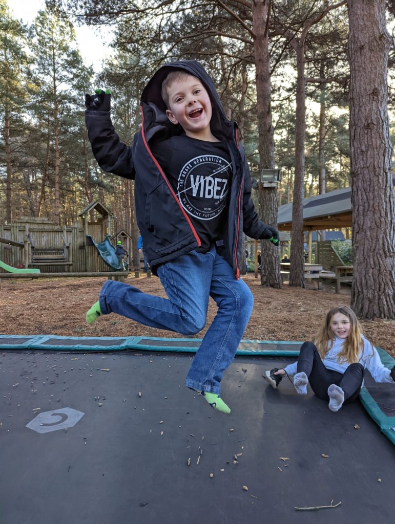 a small boy in a raincoat jumping on a trampoline with a pine forest behind