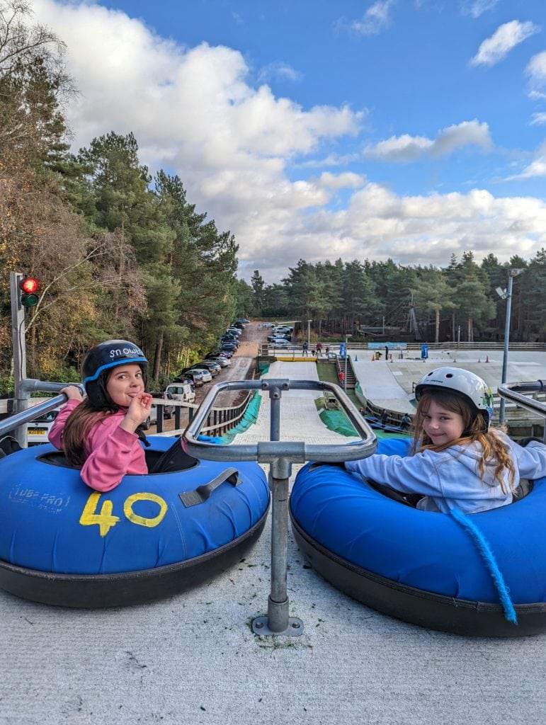 two young girls sitting in blue rubber rings at the top of a dry ski slope with green pine trees alongside it. they are turning around to look at the camera and smile. the girl on the left wears a pink jumper and a blue helmet and the girl on the right is wearing a grey jumper and a white helmet. 