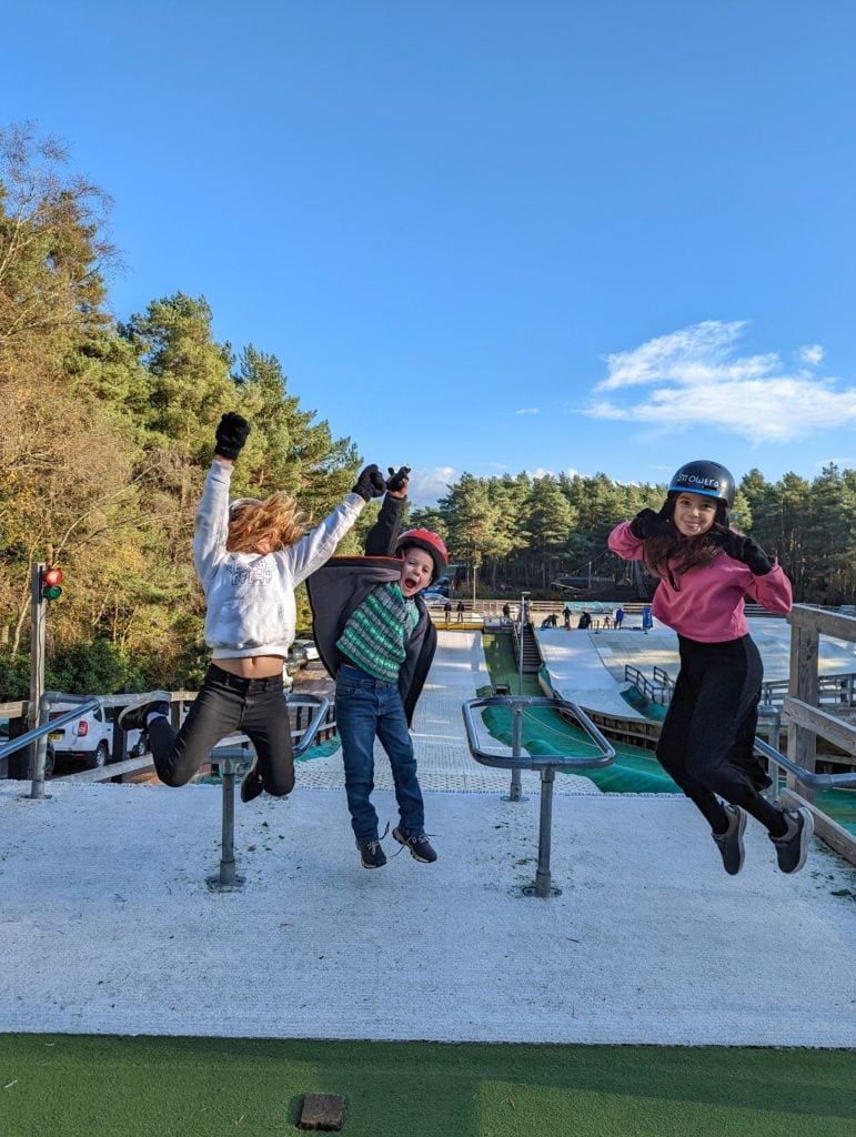 3 young kids at the top of a dry ski slope at Snowtrax Bournemouth jumping into the air under a blue sky