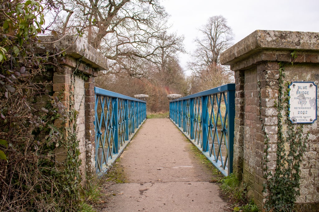looking along a stone bridge with blue metal railings otowards bare trees and a grey sky