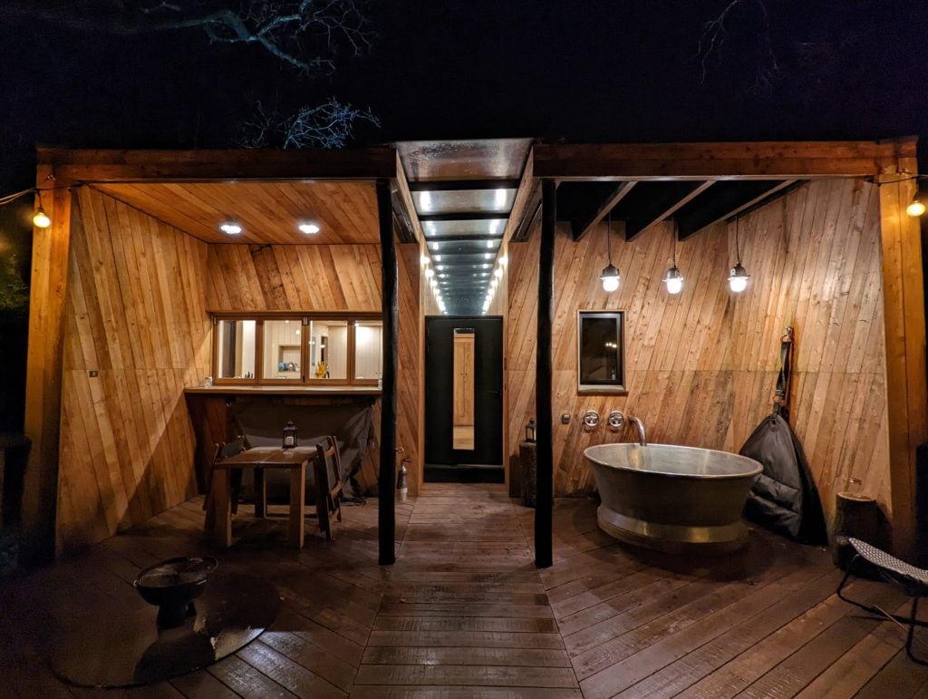 wet wooden deck of a dorset treehouse at night fter rain with electric lights and a tin hot tub 