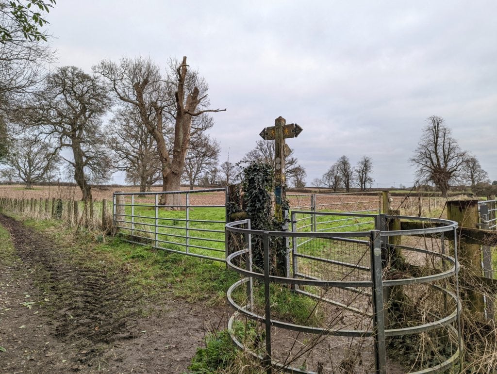 a muddy path on a dorchester river walk with a metal gate and wooden signpost with bare trees and a grey sky behind