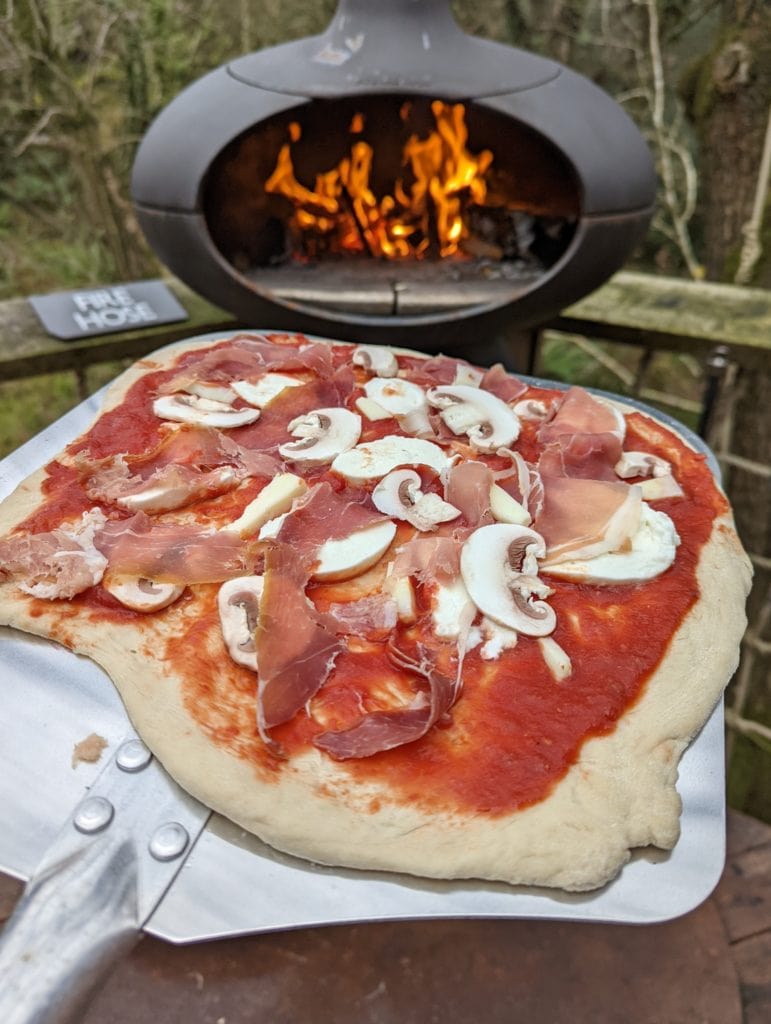 close up of a pizza with ham and mushrooms in front of a wood fired pizza oven