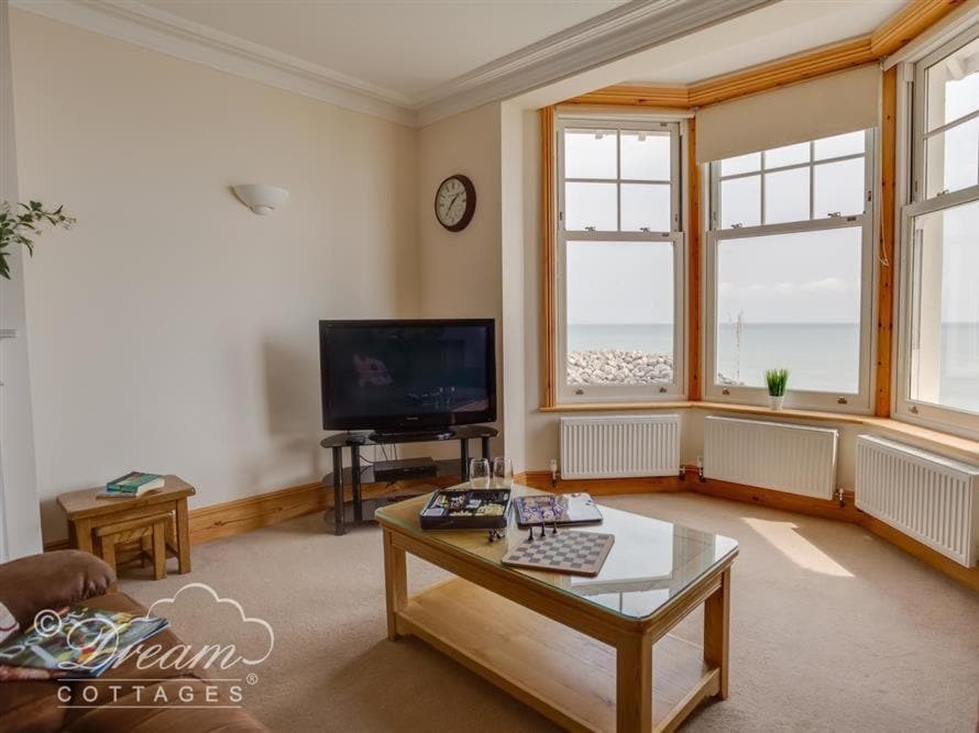 a living room with a tv, wooden coffee table, and cream walls and carpet with a large bay window overlooking the sea  - best holiday homes in weymouth