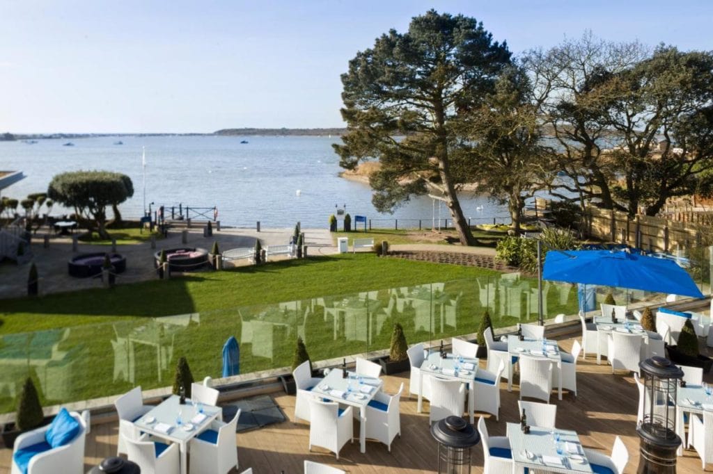 looking down at a restaurant terrace with many white tables and chairs in front of a lawn with the sea behind