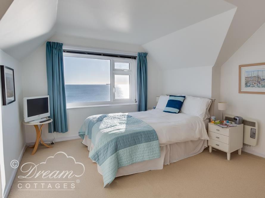 a white bedroom with a large window overlooking the sea, with blue curtains and a double bed with white bedding and a blue throw