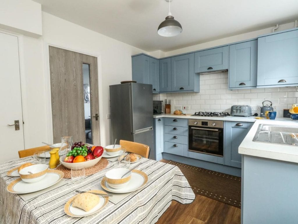 a kitchen table laid with plates and a bowl of fruit. the kitchen has blue cupboards and a silver fridge. 