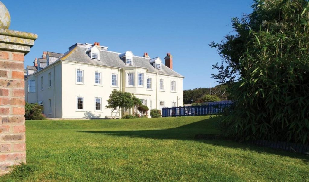 a two storey white manor house with a grey slate roof on a lawn with a blue sky behind and some trees in the foreground - moonfleet manor is one of the best spa hotels in dorset