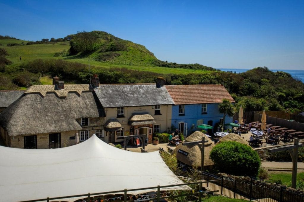 a marquee in front of 3 terraced cottages with thatched roofs. there are green grassy cliffs behind and a patch of sea visible in the distance