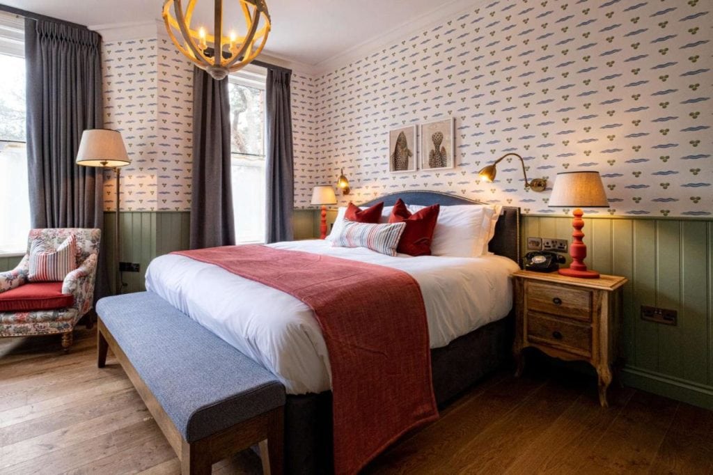 neatly made hotel bed with white sheets and a red throw and colourful patterned wallpaper behind