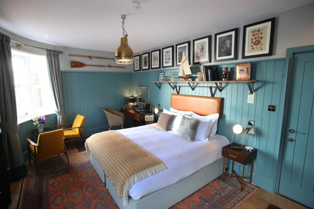 a hotel bedroom with blue painted wooden panels on the walls and lots of pictures in frames. the bed is neatly made with white sheets and an orange throw. 
